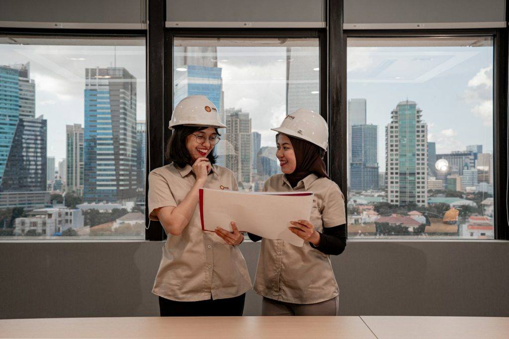 Two women in hard hats reviewing project documents beside a window