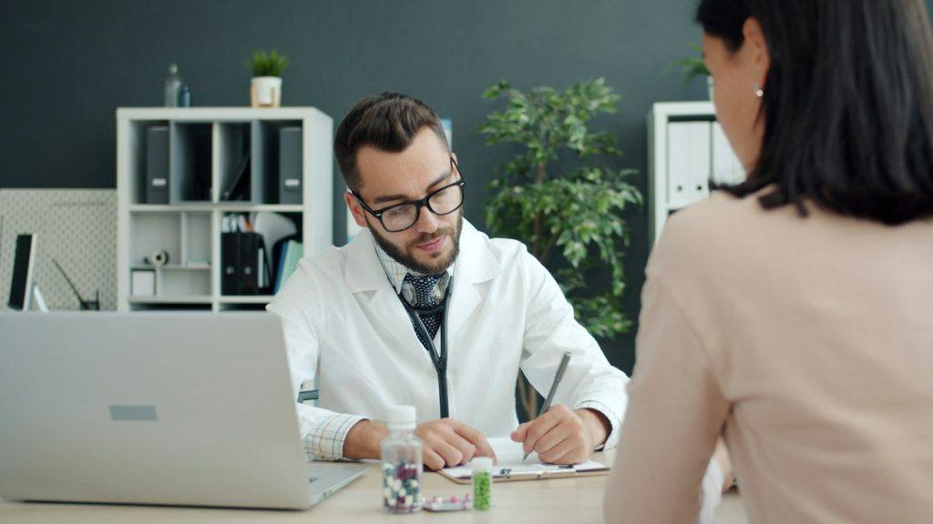 Doctor writing medical notes while a patient sits across the desk