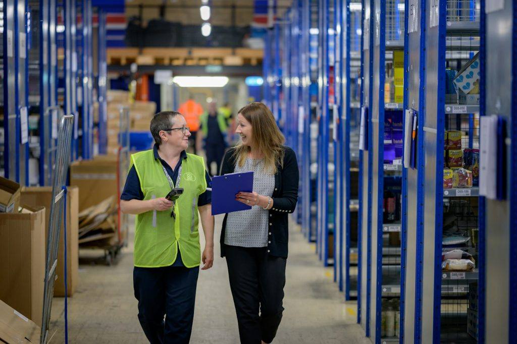 Two women with a clipboard walking down a stocked warehouse aisle